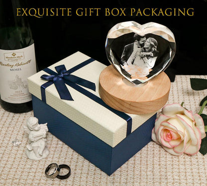 A heart-shaped crystal block featuring a black and white photograph of a couple, mounted on a round wooden base. The set includes a cream and navy blue gift box with a ribbon bow, a wine bottle, a small cherub figurine, and two rings.