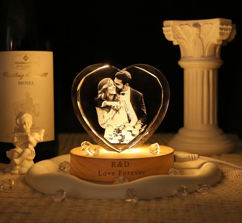 A heart-shaped glass block featuring a laser-etched photograph of a couple, illuminated by a round wooden base. The base is engraved with the text R&D and Love Forever, and is surrounded by small clear crystals on a white tray.