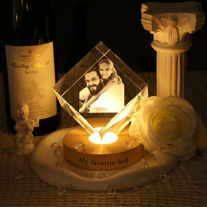 A diamond-shaped glass block featuring a laser-etched photograph of a man and a child, resting on a circular wooden base illuminated from below. The base is inscribed with the text My favorite dad in a dark, sans-serif font.