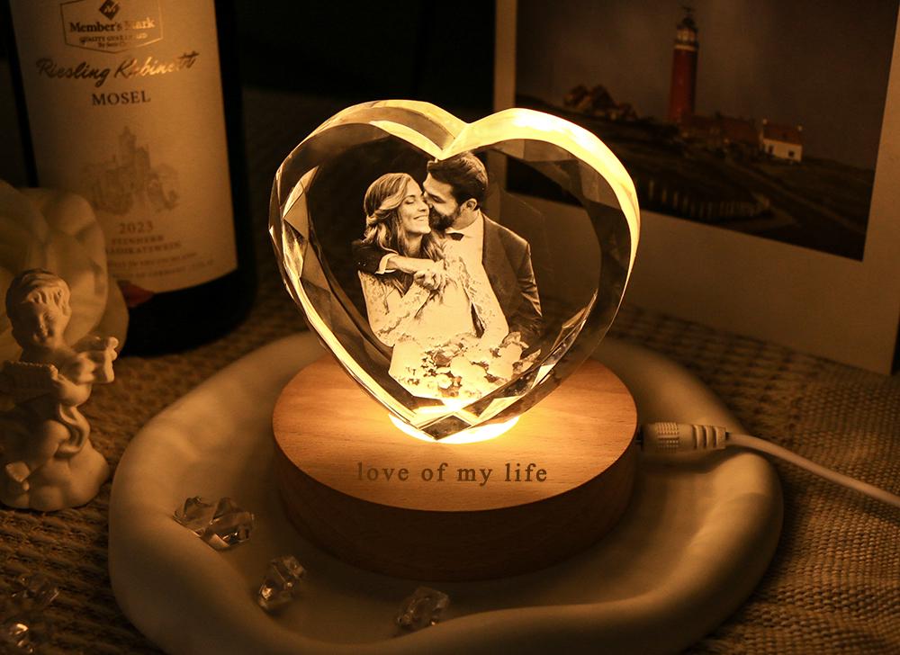 A heart-shaped crystal block featuring a laser-etched photograph of a couple, illuminated from below by a round wooden base. The base is engraved with the text "love of my life" and includes a white power cord extending to the right side.