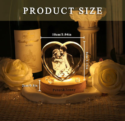 A heart-shaped crystal block featuring an engraved photograph of a couple, mounted on a round wooden base with the text "Peter & Jenny" printed on the front. The crystal measures 3.94 inches wide by 3.15 inches high, with a 0.8 inch base.