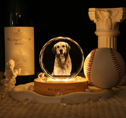 A circular crystal plaque featuring a laser-etched portrait of a golden retriever, mounted on a round wooden base illuminated from below. The base is engraved with the text "My pet : Jimmy" and sits near a baseball and wine bottle.