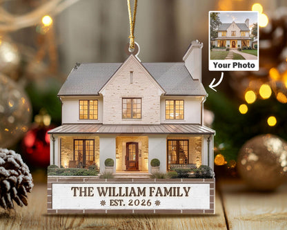 May include: A custom house-shaped Christmas ornament featuring a two-story home with a white brick facade, gray roof, and warm interior lighting. The base includes the text THE WILLIAM FAMILY and EST. 2026, with a gold hanging cord attached.