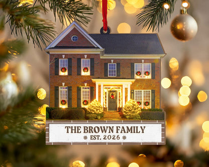 May include: A two-story brick house Christmas ornament with dark shutters, illuminated windows featuring wreaths, and a front porch with glowing lights. A white rectangular base displays the text THE BROWN FAMILY EST. 2026 with snowflake icons.