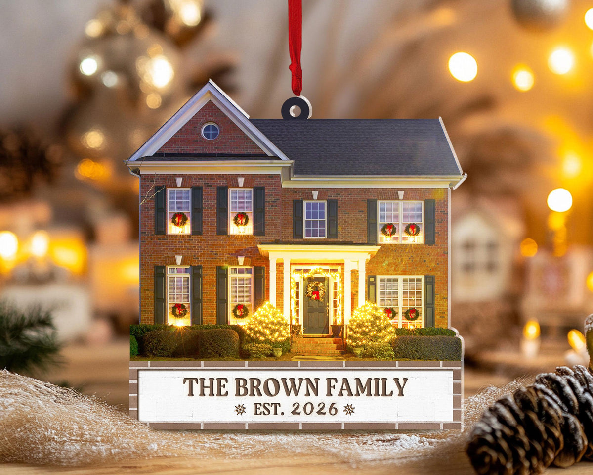 May include: A flat, house-shaped Christmas ornament featuring a two-story brick home with dark shutters, illuminated windows with wreaths, and a front porch. A white base displays the text THE BROWN FAMILY and EST. 2026 with a red ribbon for hanging.