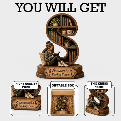 A brown, S-shaped bookshelf figurine featuring a person reading a book, with shelves holding miniature books, plants, and a mug. A plaque reads 'Samantha's Reading Corner'. The item has a 15mm thickness and comes in a giftable box.