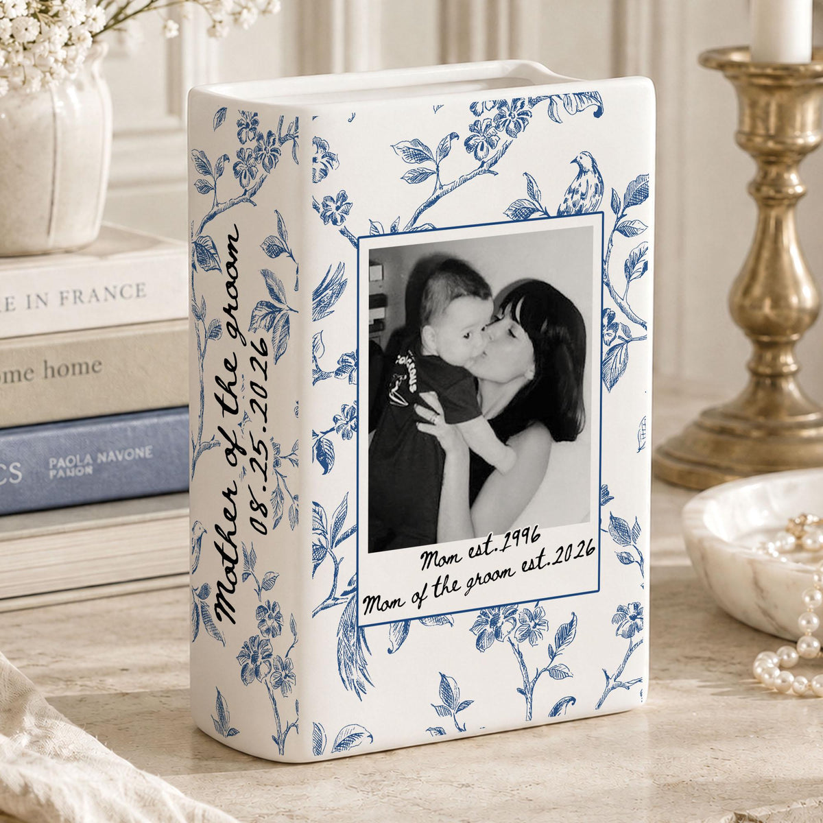 A white ceramic vase with a blue floral pattern featuring a black and white photograph of a person holding a child. The text reads Mother of the groom 08.25.2026 and Mom est. 1996 Mom of the groom est. 2026 printed on the front surface.