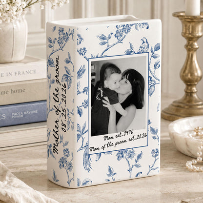 A white ceramic vase with a blue floral pattern featuring a black and white photograph of a person holding a child. The text reads Mother of the groom 08.25.2026 and Mom est. 1996 Mom of the groom est. 2026 printed on the front surface.
