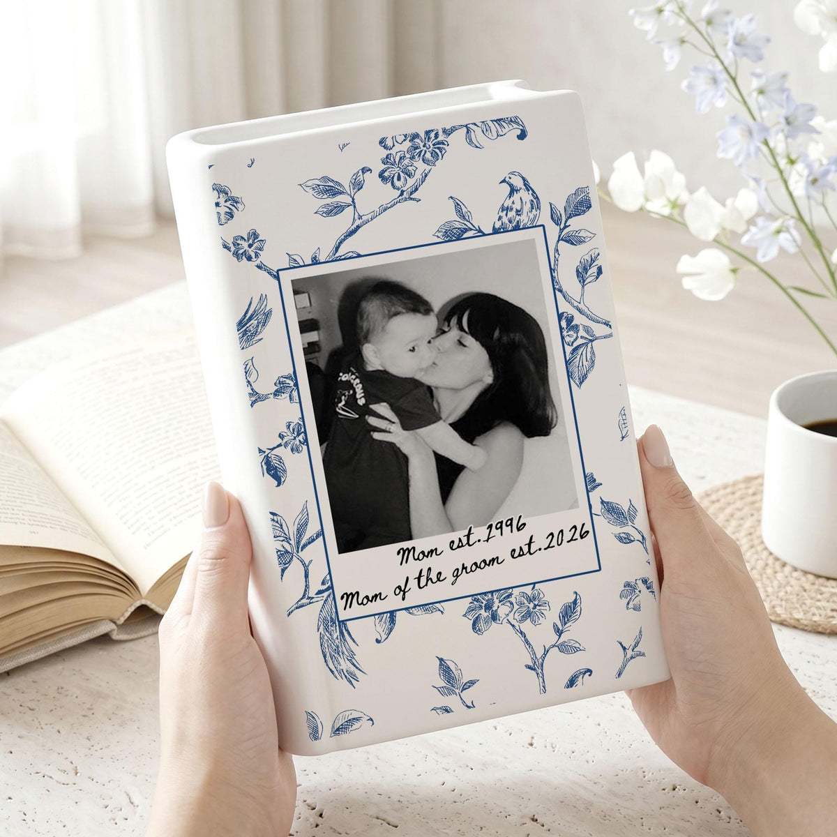 A white ceramic vase with a blue floral pattern featuring a central rectangular frame containing a black and white photograph of a person holding a baby. The text "Mom est. 1996" and "Mom of the groom est. 2026" is printed below the photo.