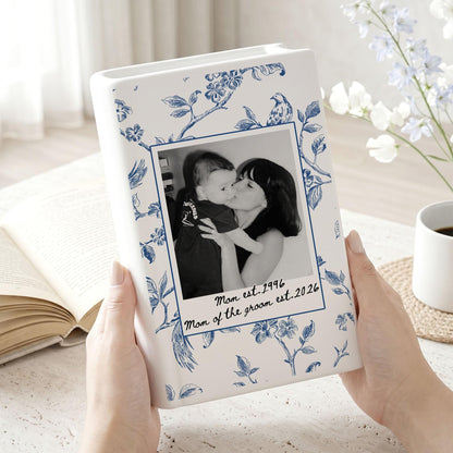 A white ceramic vase with a blue floral pattern featuring a central rectangular frame containing a black and white photograph of a person holding a baby. The text "Mom est. 1996" and "Mom of the groom est. 2026" is printed below the photo.