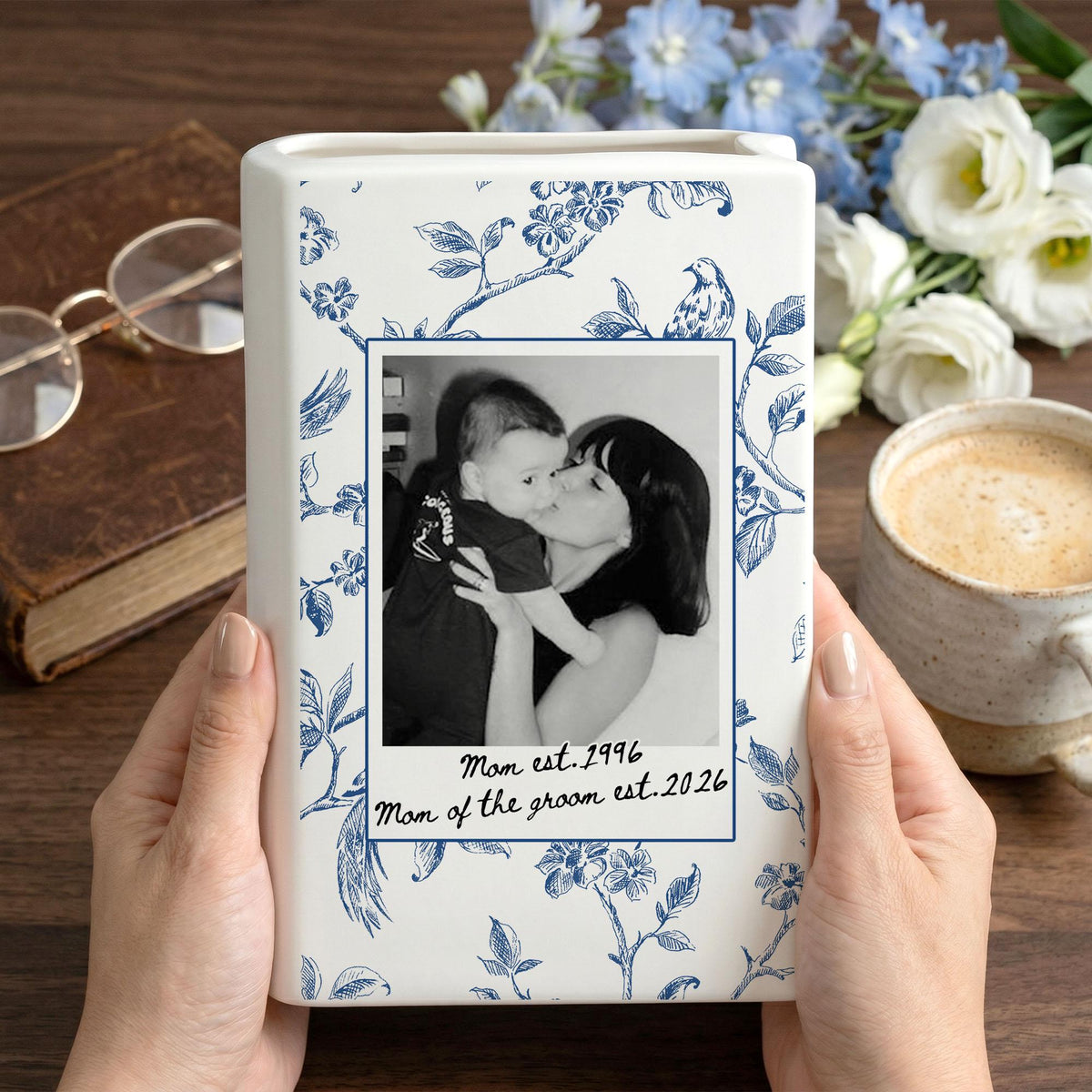 A white ceramic rectangular vase featuring a blue floral pattern and a central black and white photograph of a person holding a baby. The text below the photo reads: Mom est. 1996, Mom of the groom est. 2026, framed in a blue border.