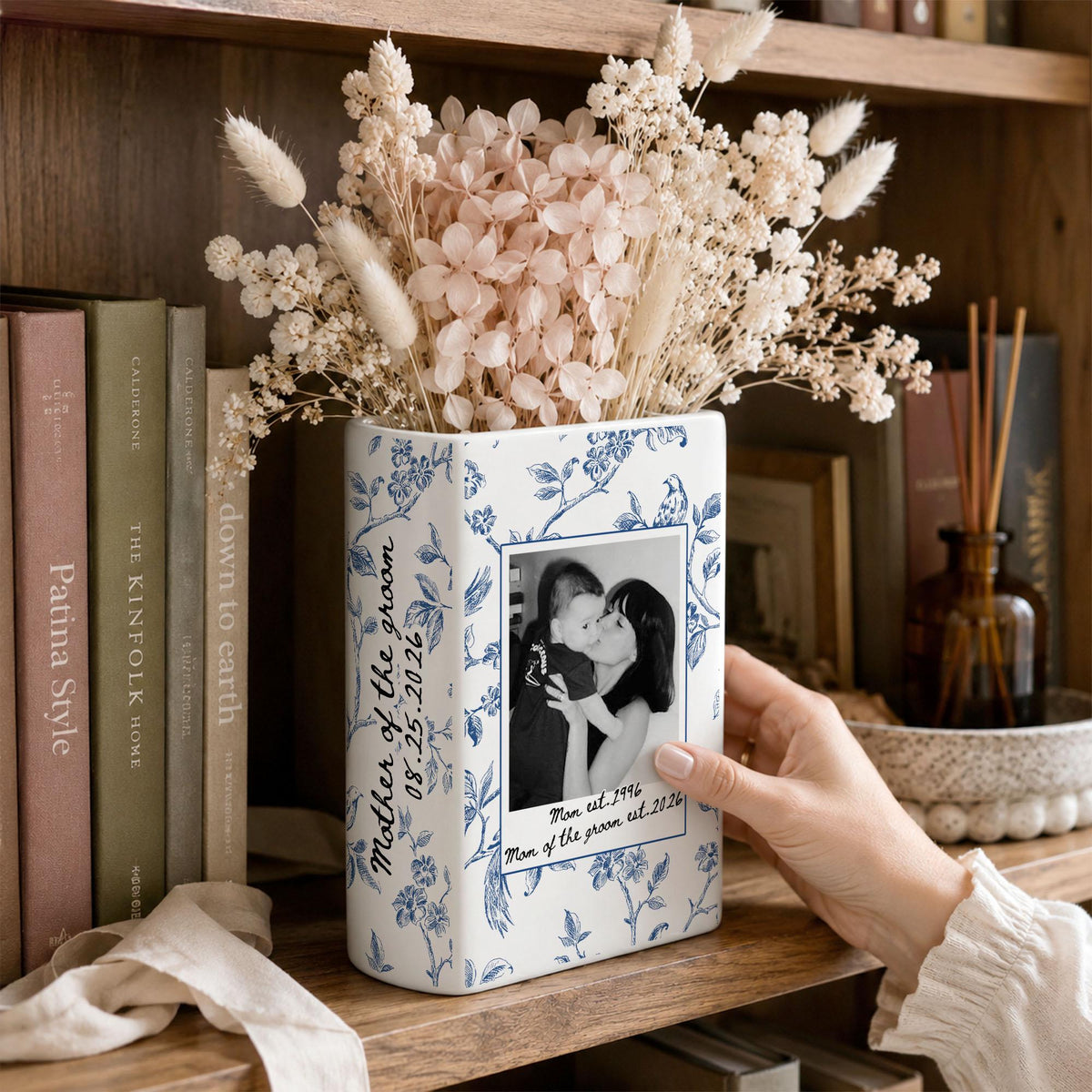 A white ceramic book-shaped vase featuring a blue floral pattern. The front displays a black and white photograph of two people with the text Mother of the groom 08.25.2026 and Man of the groom est. 1996 Man of the groom est. 2026.