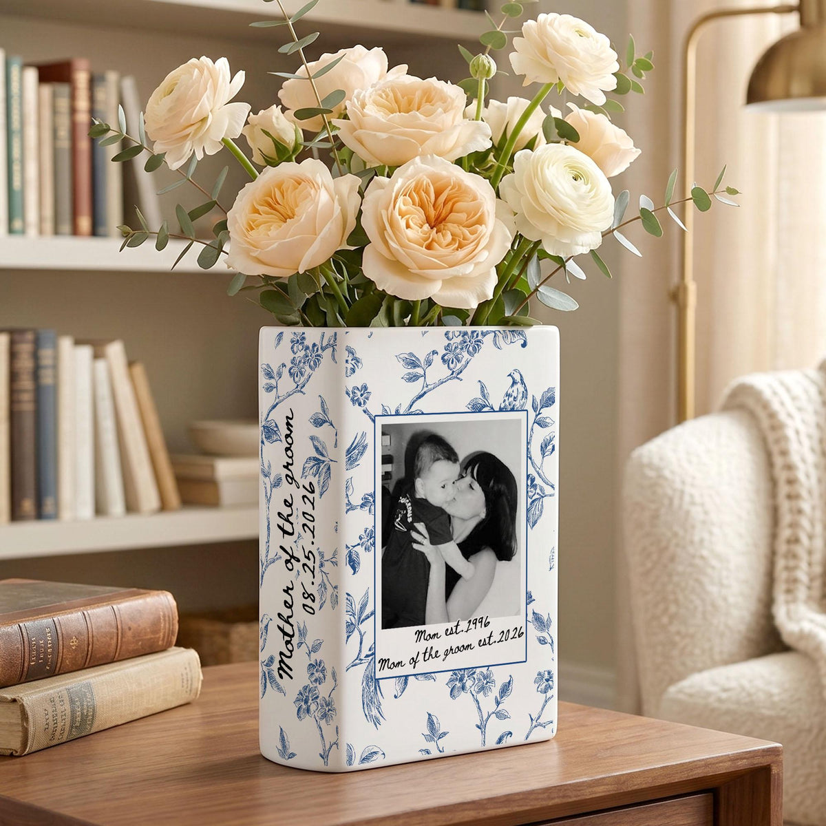 A rectangular white ceramic vase with a blue floral pattern, featuring a black and white photograph of a person holding a child. The text reads Mother of the groom 08.25.2026 and Man est. 1996 Mom of the groom est. 2026. Filled with pale peach flowers.