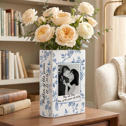 A rectangular white ceramic vase with a blue floral pattern, featuring a black and white photograph of a person holding a child. The text reads Mother of the groom 08.25.2026 and Man est. 1996 Mom of the groom est. 2026. Filled with pale peach flowers.