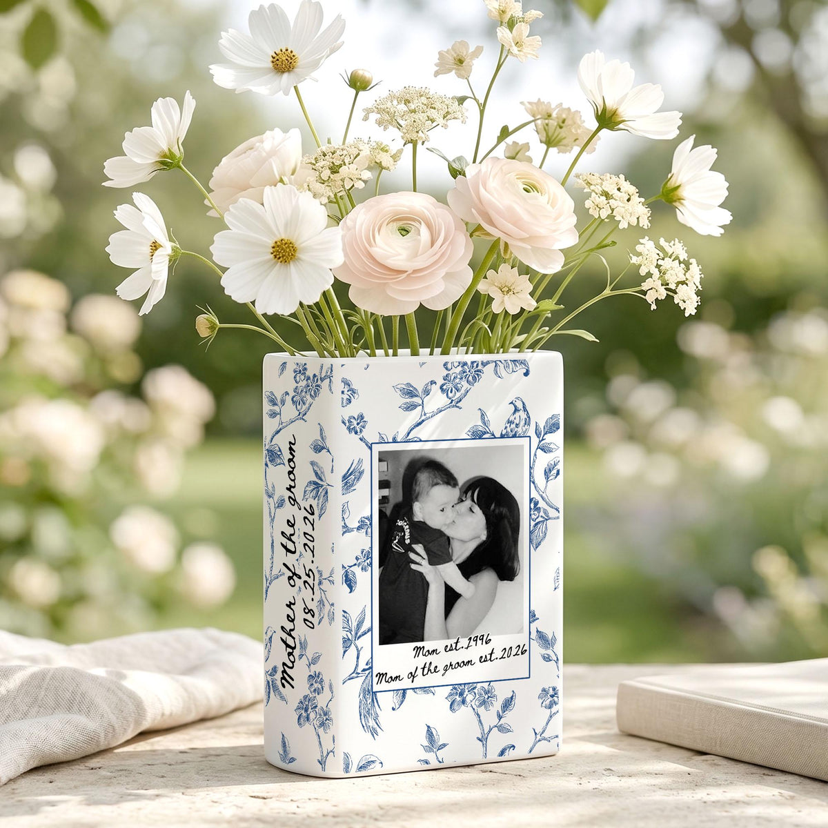 A rectangular white ceramic vase with a blue floral pattern, filled with white cosmos and ranunculus flowers. The vase features a black and white photograph of a person holding a child, with the text "Mother of the groom 08.25.2026" and "Mom est. 1996 Mom of the groom est. 2026" printed on the front.
