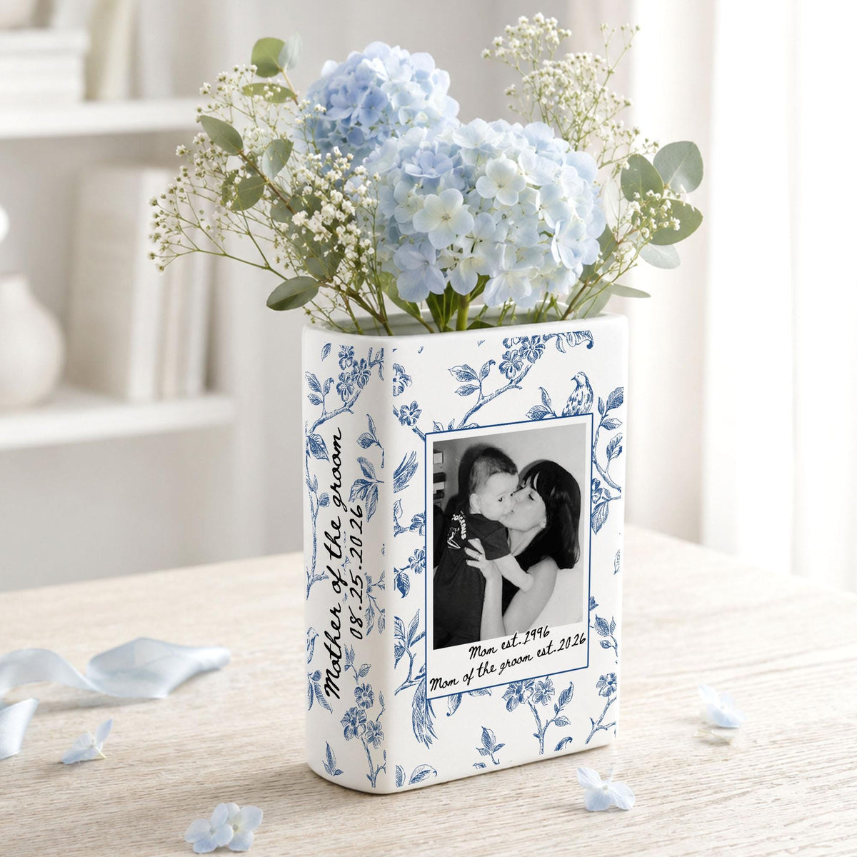 A rectangular white ceramic vase featuring a blue floral pattern, a black and white photograph of two people, and the text "Mother of the groom 08.25.2026" and "Mom est. 1996 Mom of the groom est. 2026" filled with light blue hydrangea flowers.