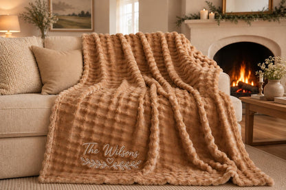 A tan-colored, textured throw blanket with a waffle-like pattern draped over a sofa. The blanket features white embroidered text that reads The Wilsons, positioned above a small heart icon and a decorative leafy branch design.