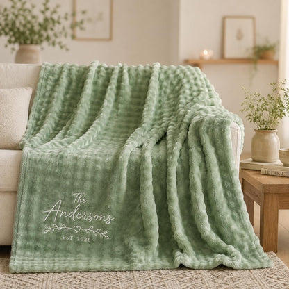 A sage green textured throw blanket featuring a soft, dimpled surface. The blanket is personalized with white embroidered text that reads The Andersons, accompanied by a small heart and leaf design, with the text EST. 2026 below it.