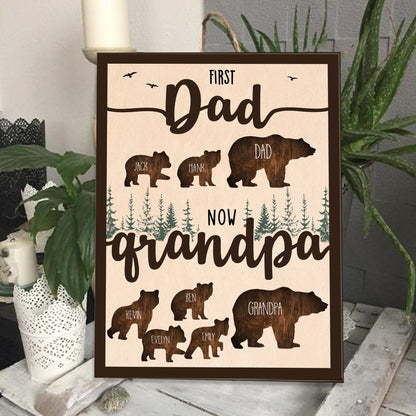 May include: A brown wooden sign with the text "First Dad Now Grandpa" in brown lettering. The sign features a silhouette of a bear family with the names "Jack", "Hank", "Dad", "Kevin", "Ben", "Grandpa", "Evelyn", and "Emily" written below each bear.