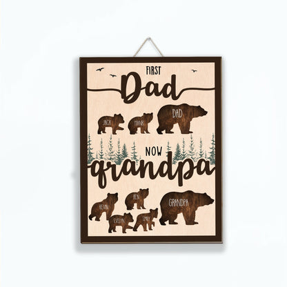 May include: A wooden sign with a brown background and a light brown wood grain texture. The sign features a silhouette of a bear family with the text "First Dad Now Grandpa" and the names of the family members.