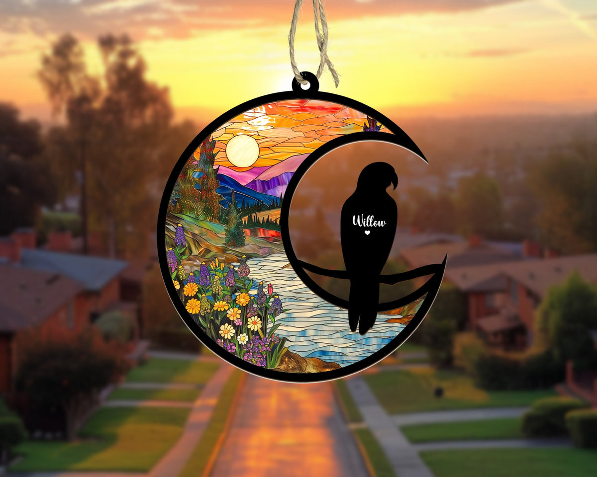 May include: A stained glass style ornament featuring a crescent moon with a silhouette of a bird perched on a branch. The background shows a colorful landscape with mountains, a river, and flowers. The bird has the name "Willow" written on it.