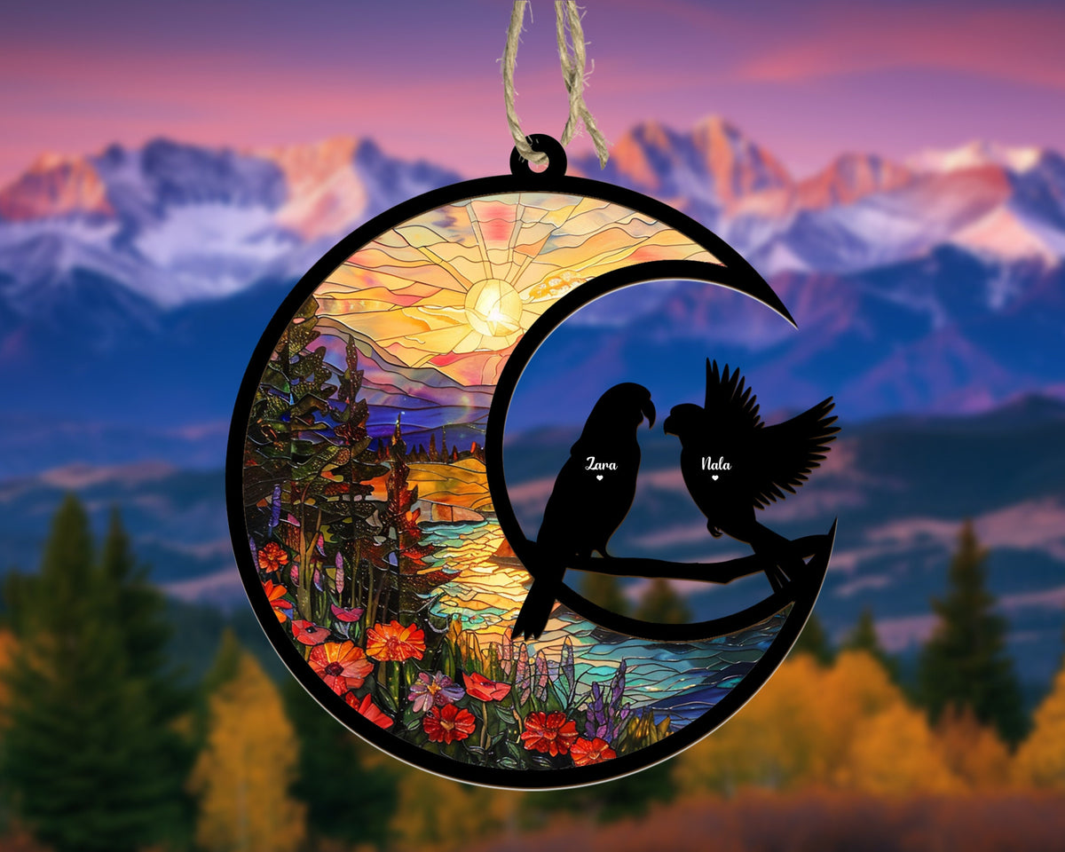 May include: A stained glass ornament shaped like a crescent moon with a sunrise scene and two birds perched on a branch. The birds are silhouetted and have the names "Zara" and "Isla" written on them.