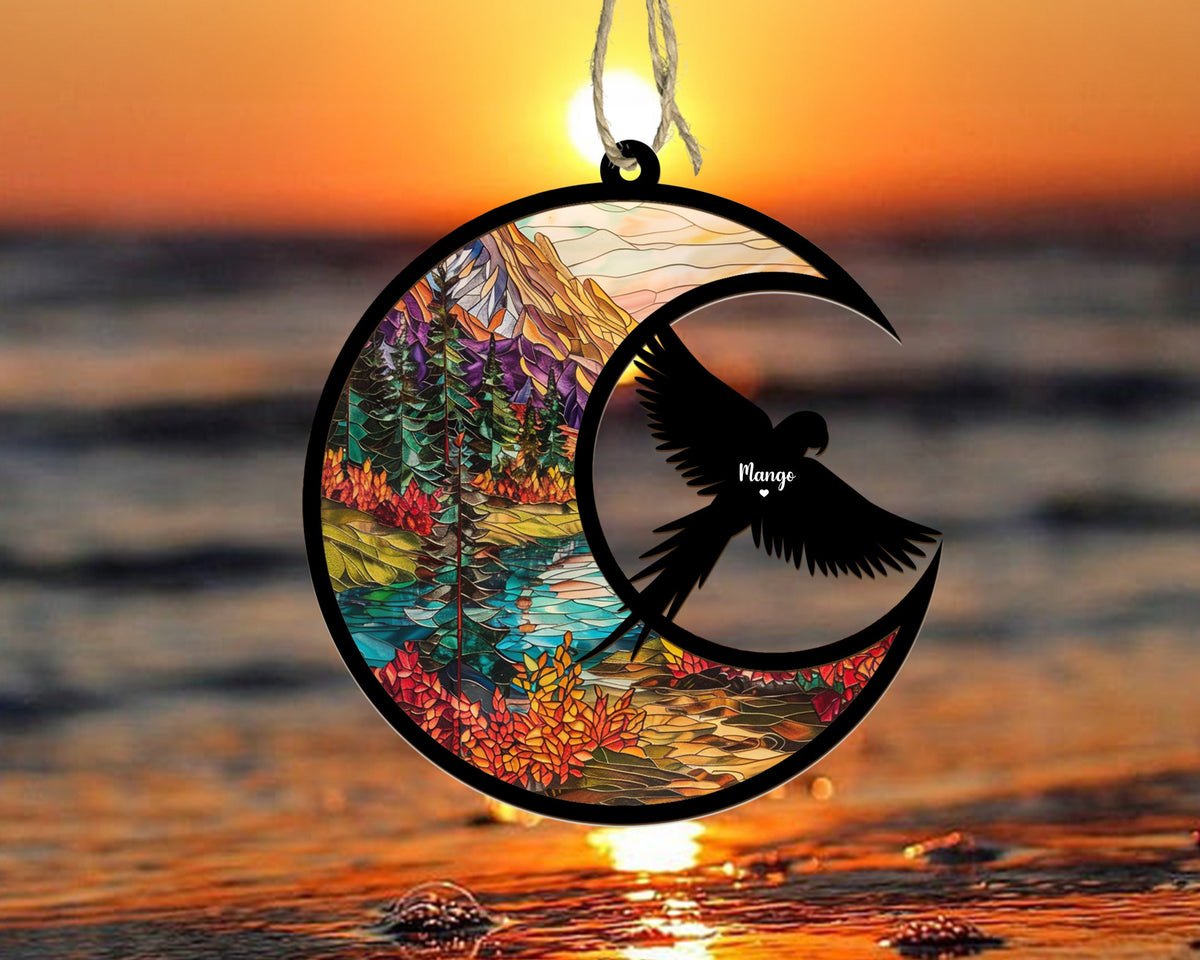 May include: A stained glass ornament shaped like a crescent moon with a silhouette of a bird flying inside. The moon features a colorful landscape with trees, mountains, and a river. The bird has the name "Mango" written on it.