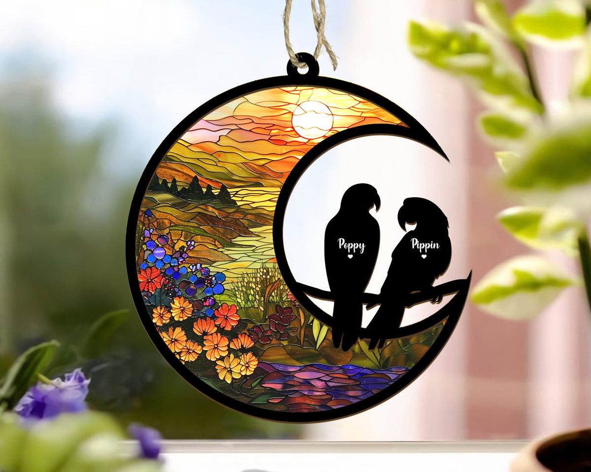 May include: A stained glass style moon ornament with a sunset scene and two black silhouettes of parrots perched on a branch. The parrots are labeled "Peppy" and "Pippin".