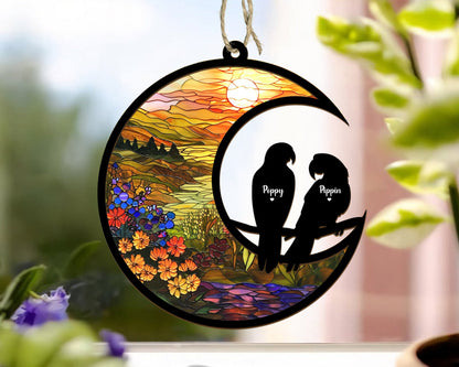 May include: A stained glass style moon ornament with a sunset scene and two black silhouettes of parrots perched on a branch. The parrots are labeled "Peppy" and "Pippin".