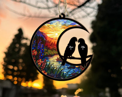May include: A wooden ornament shaped like a crescent moon with a stained glass design of a sunset over a lake and mountains. Two black silhouettes of birds are perched on a branch inside the moon, with the names "Kiwi" and "Mike" written on them.