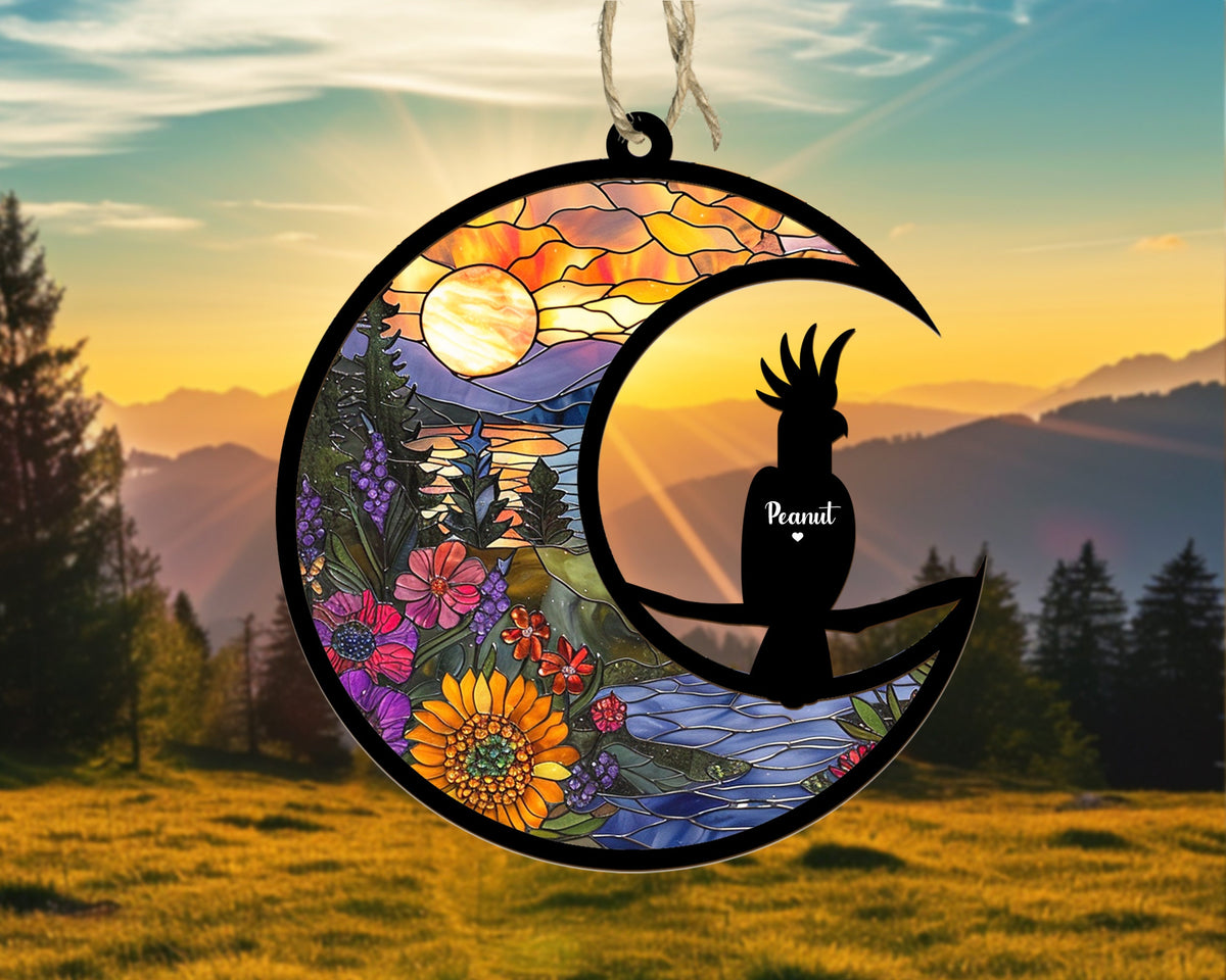 May include: A stained glass style ornament featuring a crescent moon with a sunset scene and a black silhouette of a cockatoo bird perched on a branch. The bird is labeled "Peanut" with a heart symbol.