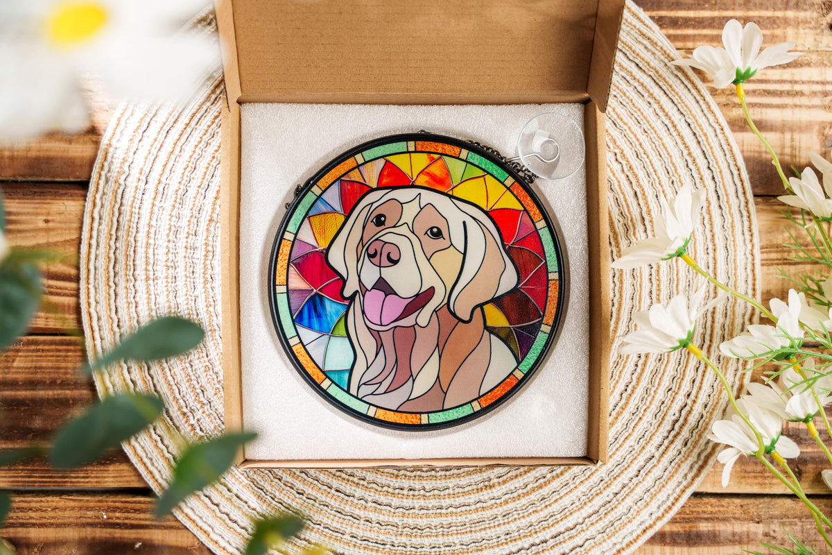 May include: A stained glass window hanging featuring a yellow Labrador Retriever with a colorful background. The dog is looking directly at the viewer with its mouth open and tongue sticking out. The background is a mix of red, orange, yellow, green, blue, and purple.