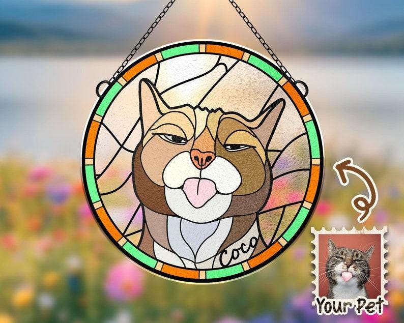 May include: A stained glass window hanging with a cat's face. The cat has brown fur, green eyes, and a pink tongue sticking out. The cat's name is "Coco" written on the glass. The window is surrounded by a green, orange, and yellow frame.