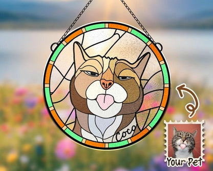 May include: A stained glass window hanging with a cat's face. The cat has brown fur, green eyes, and a pink tongue sticking out. The cat's name is "Coco" written on the glass. The window is surrounded by a green, orange, and yellow frame.