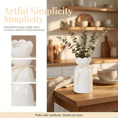 A white ceramic vase with a scalloped rim and a smooth, high-gloss finish. The neck of the vase is tied with a thin, cream-colored ribbon. The vase contains a sprig of dried eucalyptus leaves and is displayed on a wooden surface.
