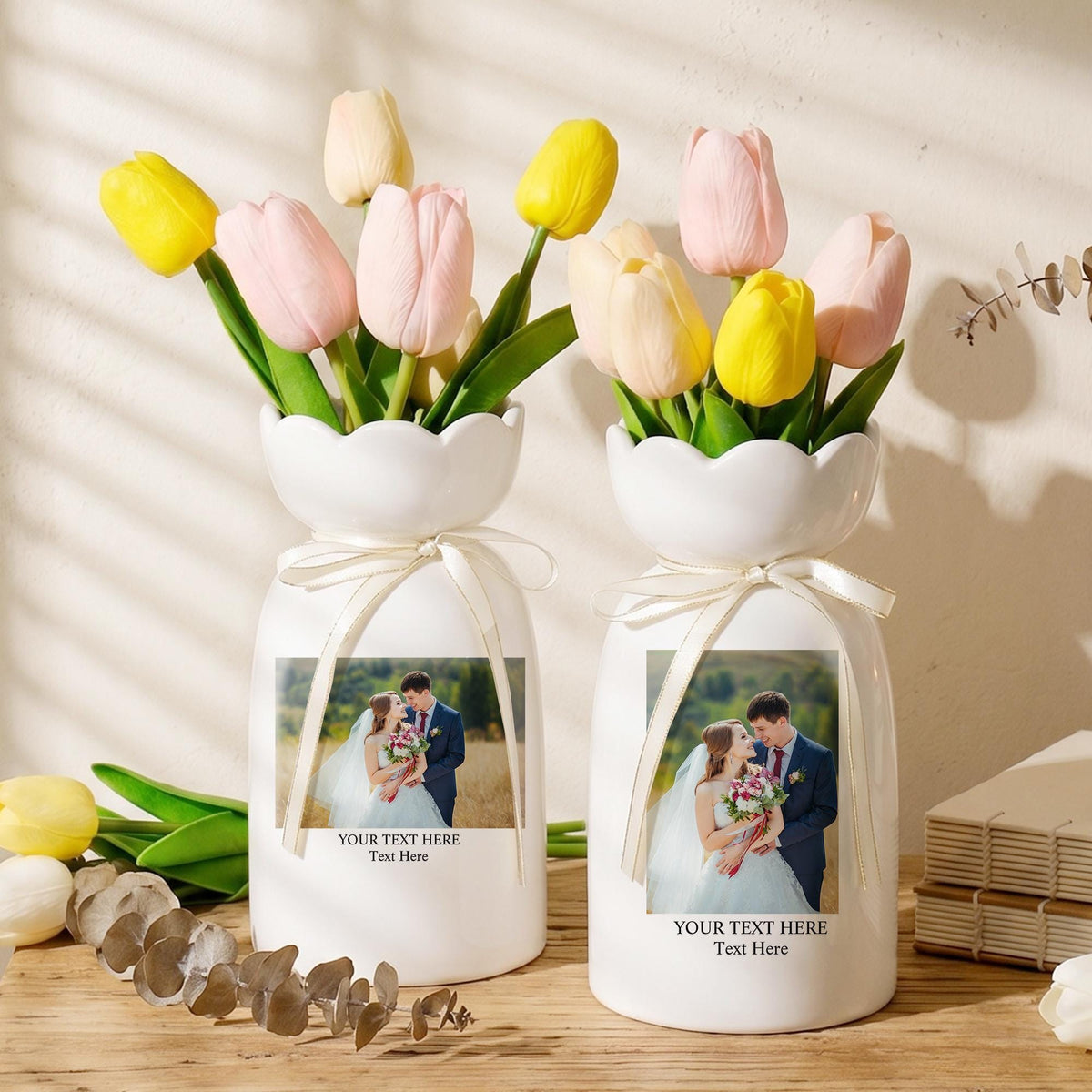 Two white ceramic vases with scalloped rims, each tied with a cream ribbon. The vases feature a printed photograph of a couple in wedding attire and the text "YOUR TEXT HERE Text Here". Each vase holds a bouquet of yellow and pink tulips.
