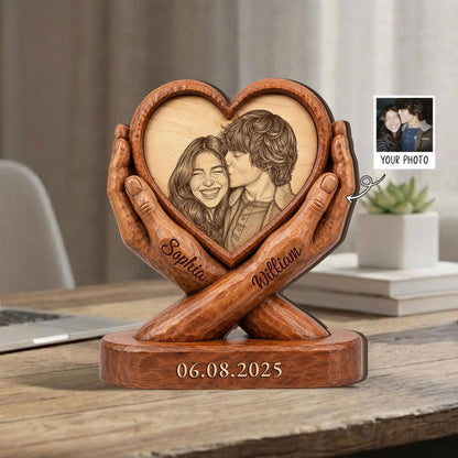 A wooden heart-shaped photo frame held by carved hands. The frame features a laser-etched image of a couple and is personalized with the names "Sophia" and "William." The base displays the date "06.08.2025."