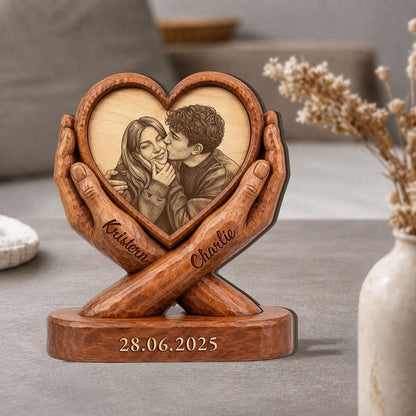 A wooden heart-shaped photo frame held by carved hands. The frame contains a black and white image of a couple. The names "Kristen" and "Charlie" are engraved on the hands, and the date "28.06.2025" is on the base.