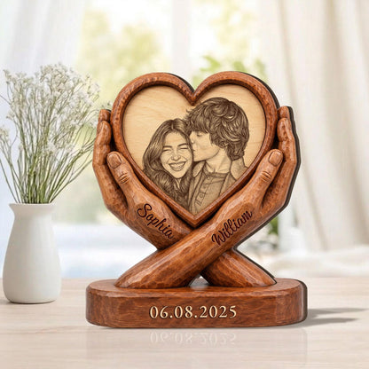 A wooden heart-shaped photo frame with a carved image of a couple. The frame is held by carved wooden hands. The names "Sophia" and "William" are carved on the hands. The date "06.08.2025" is carved on the base.