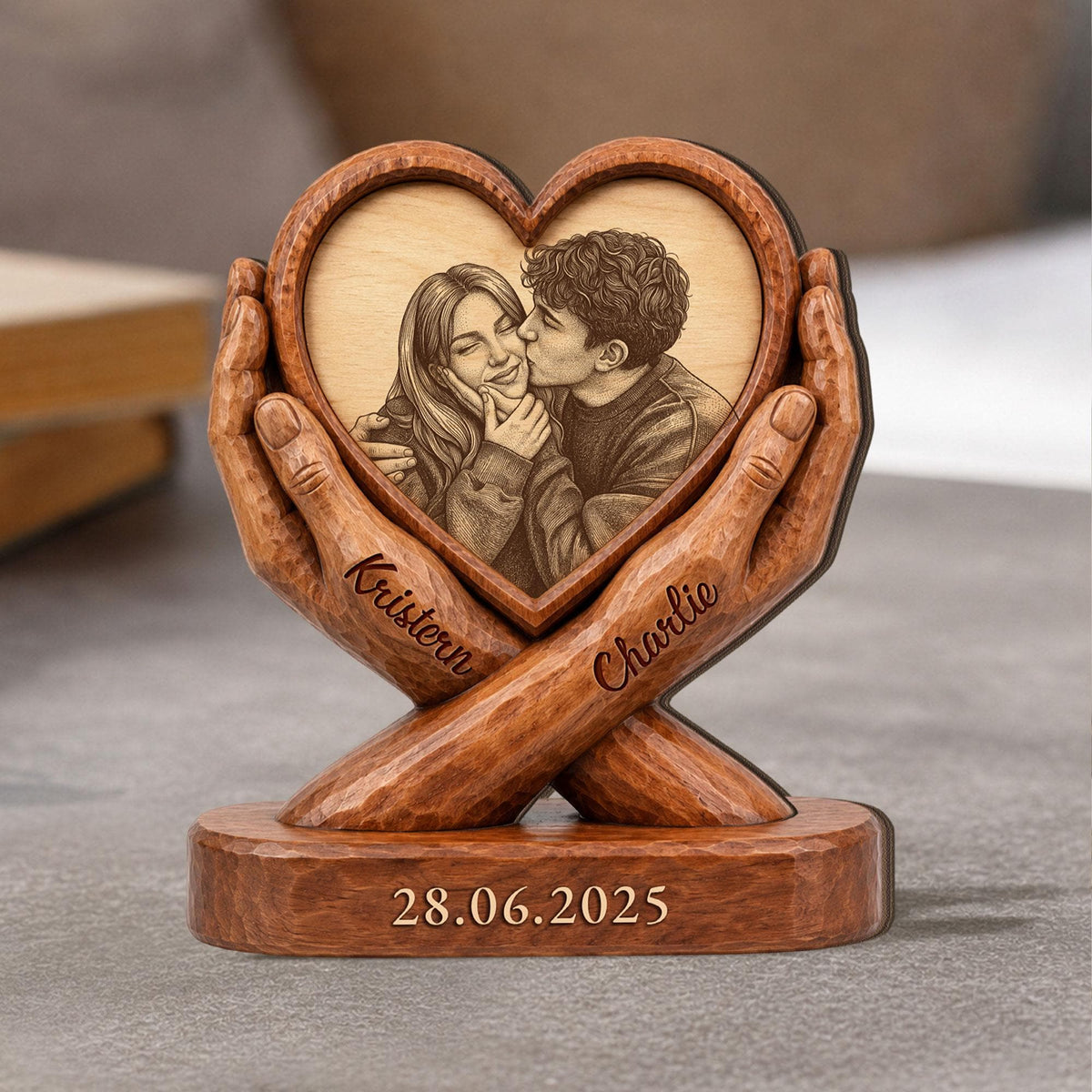 A wooden heart-shaped photo frame with a carved image of a couple kissing. The frame is held by carved wooden hands. The names "Kristen" and "Charlie" are inscribed on the hands, and the date "28.06.2025" is at the base.