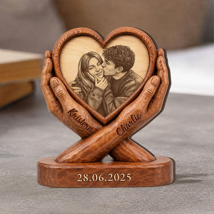 A wooden heart-shaped photo frame with a carved image of a couple kissing. The frame is held by carved wooden hands. The names "Kristen" and "Charlie" are inscribed on the hands, and the date "28.06.2025" is at the base.