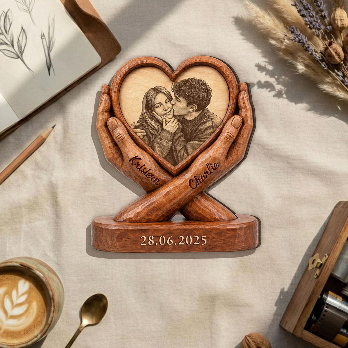A wooden heart-shaped photo frame with a couple's image, held by carved hands. The names "Kristiern" and "Charlie" are inscribed on the hands. The base displays the date "28.06.2025".