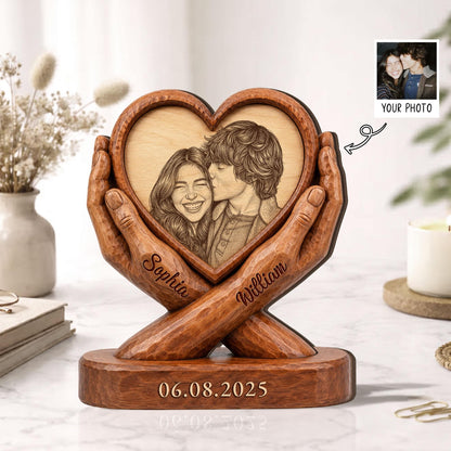 A wooden heart-shaped photo frame held by carved hands. The frame contains a black and white portrait of a couple. The names "Sophia" and "William" are inscribed on the hands. The date "06.08.2025" is engraved on the base.