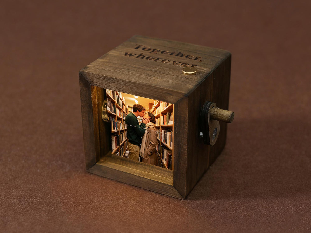 A wooden cube-shaped photo box with the words "together, wherever" engraved on top. The box features a small photo of a couple kissing inside a library. A small, gold-colored ring is on top.