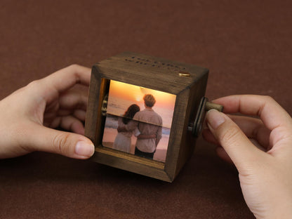 A wooden photo cube with a rotating mechanism, displaying a couple on a beach at sunset. The cube is dark brown with a small handle and a gold-colored accent. The image is illuminated from within.