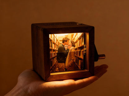 A wooden box with a rectangular window displaying a couple kissing in a library setting. The box has a crank on the side and is held in a hand. The interior scene is illuminated with warm light, creating a cozy atmosphere.