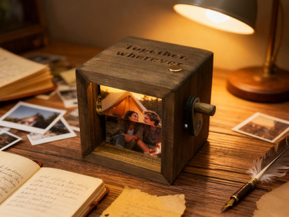 A wooden photo cube with a picture of a couple inside, illuminated by a warm light. The cube has a crank on the side and the words "Together, wherever" engraved on top. The cube sits on a wooden surface with other photos and a lamp.