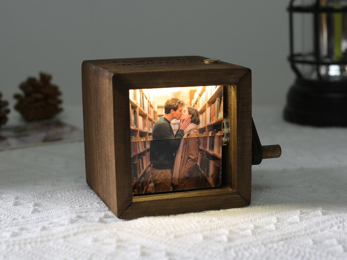 A wooden music box with a photo of a couple kissing inside. The box is square with a crank on the side and a warm light illuminating the image. The background shows a library setting.