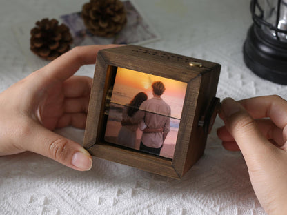 A wooden photo cube with a picture of a couple at sunset. The cube is being held by hands. The cube has a small crank on the side. The wood is a dark brown color.