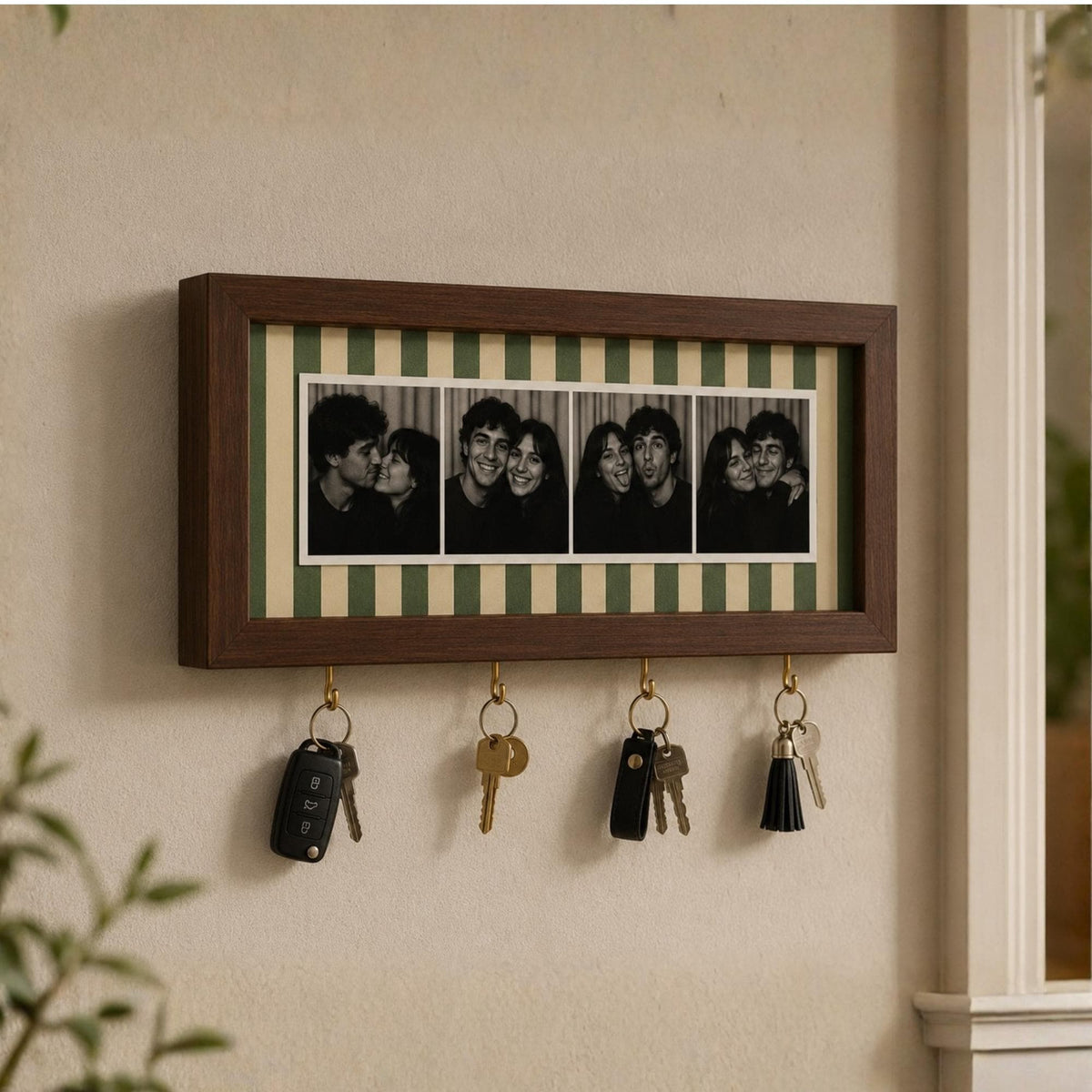 A rectangular dark wood wall-mounted key holder featuring a horizontal photo strip of four black and white portraits. The frame has a green and cream striped background and four brass-colored metal hooks holding various keys.